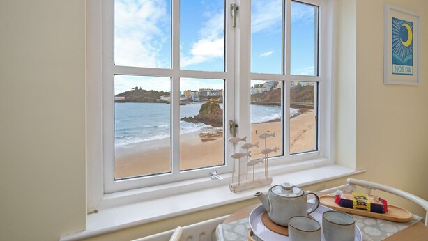 A room with a large window overlooking a beach, a small table with a teapot and cups, and a framed picture on the wall. - 5 Sunny Cove - Direct Beach Access, Sea Views (Tenby)