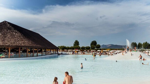 Une grande piscine extérieure, dotée d’un pavillon au toit de chaume et de parasols, entourée d’arbres et avec des montagnes en toile de fond.