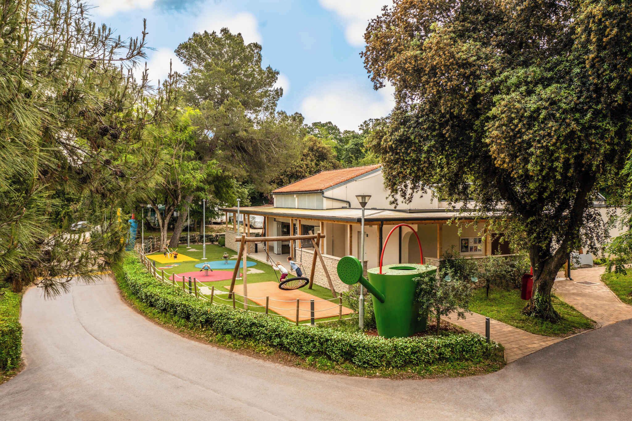 A playground with swings and a tennis court, surrounded by trees and a building with a red roof.
