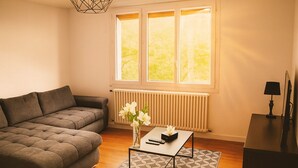 A living room with a grey sofa, a coffee table with a vase of flowers, a radiator, and a window with a view of greenery. - Maison le Ber-So Close to Beauval Zoo (Selles-sur-Cher)