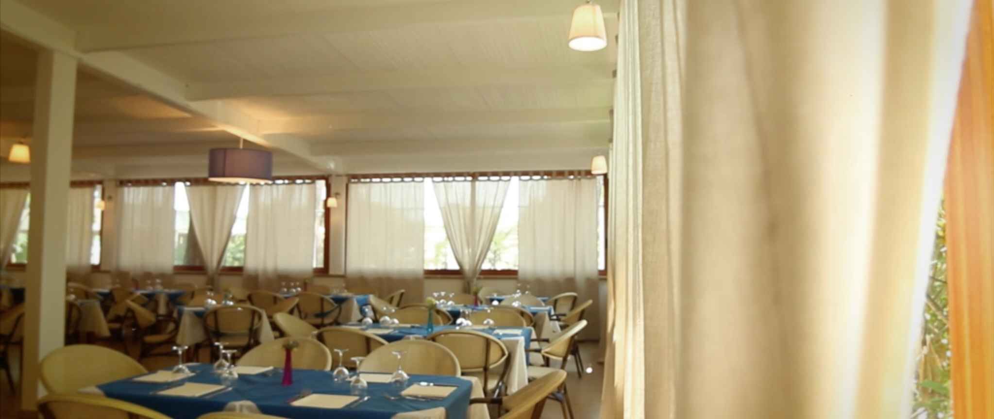 Et spiseområde med dækkede borde, store vinduer med gardiner og et loft med hængende lamper.