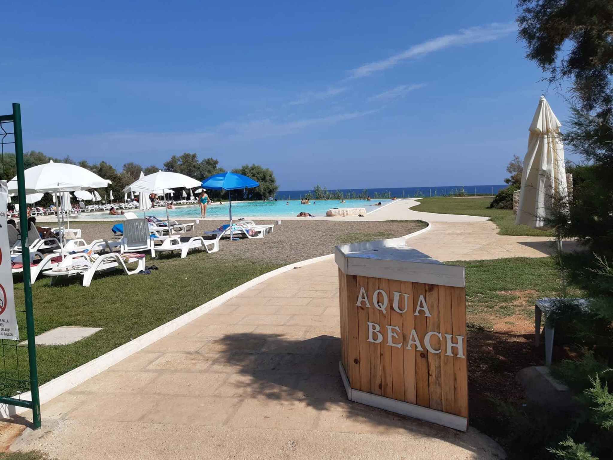 A wooden sign reading "AQUA BEACH" with a pool area, lounge chairs, and umbrellas in the background.