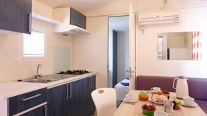 Une cuisine compacte équipée d’une cuisinière à gaz, d’un évier et de meubles modernes. On aperçoit un coin repas avec une table mise pour le petit-déjeuner, agrémenté d’une machine à café, de viennoiseries et d’un saladier de fruits.