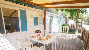 Un espace extérieur abrité comprenant une table et des chaises blanches, une terrasse en bois et une maison mobile avec des volets bleus.