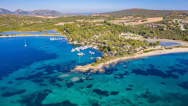 Vue aérienne d’une zone côtière avec des eaux bleues et limpides, des bateaux et un petit village composé de bâtiments et de verdure.