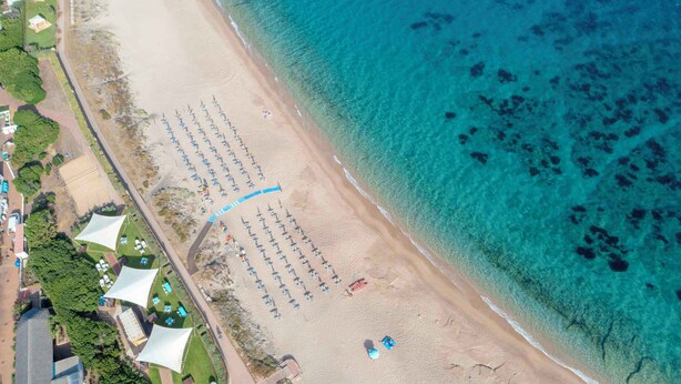 Oversigtsbillede af en strand med rækker af parasoller og liggestole, et klart blåt hav og en vej ved stranden.