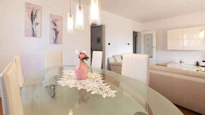 Ett modernt matområde med ett glasbord, vita stolar och en rosa vas med blommor. I bakgrunden finns väggmonterade lampor och ett vitt skåp.