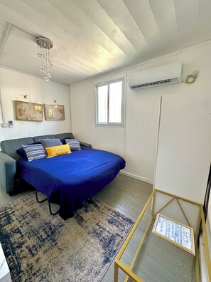 Un dormitorio con cama, sofá, candelabro, decoración en la pared, ventana y aire acondicionado.