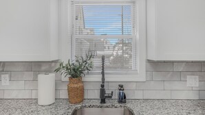 A kitchen with a stainless steel sink, a white countertop, a potted plant, and a window with blinds.