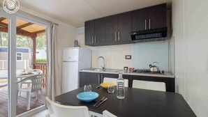 A compact kitchen with dark cabinets, a white refrigerator, a sink, and a stove. There is a dining table with chairs and a blue bowl on it. - Mobile home with air conditioning (Peschici)