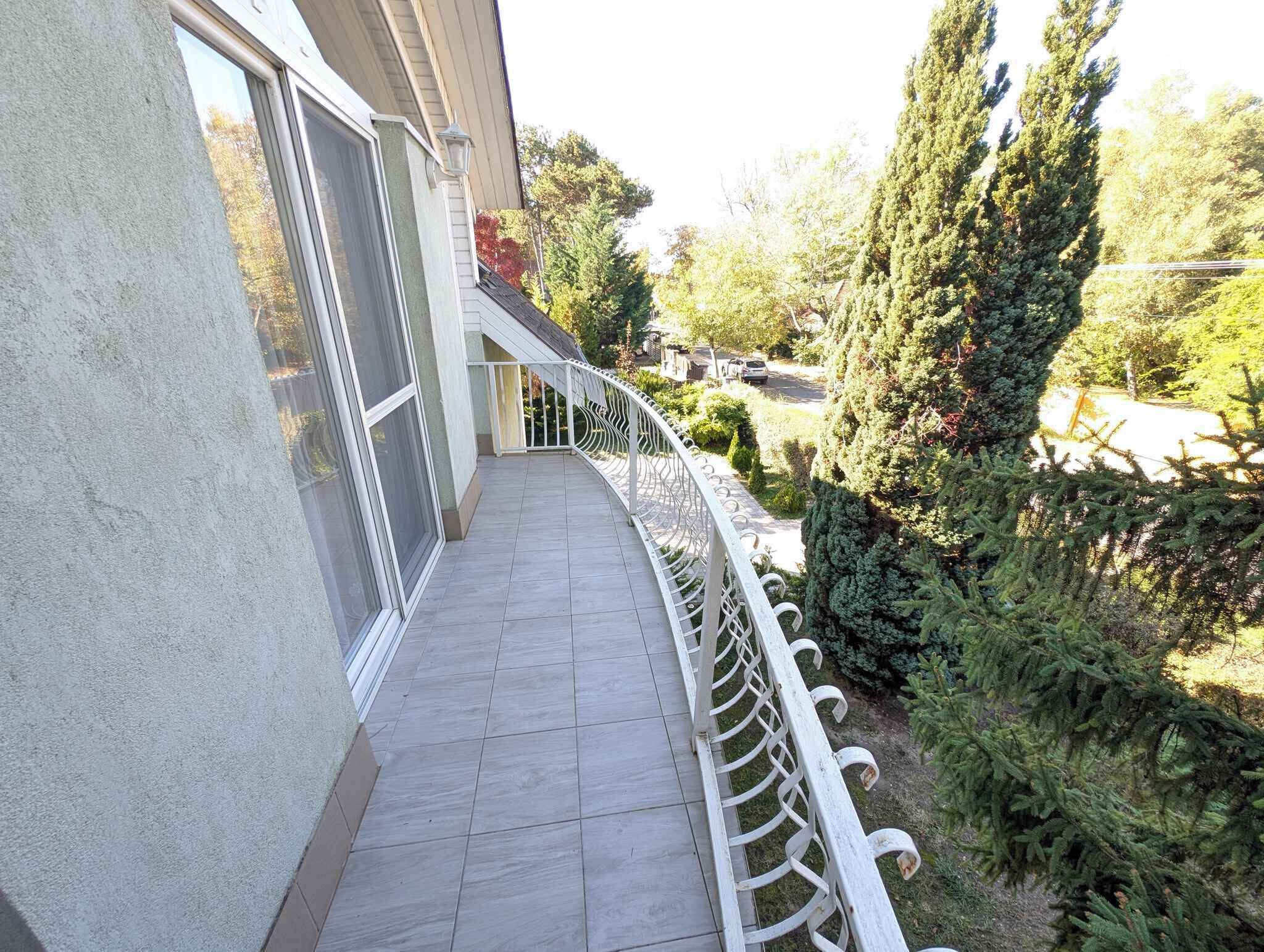 A balcony with a white railing, a tiled floor, and a view of a garden with trees and shrubs.