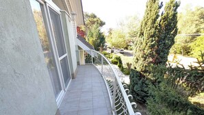 A balcony with a white railing, a tiled floor, and a view of a garden with trees and shrubs. - Holiday apartment with large balcony, American kitchen and air conditioning (Siófok)