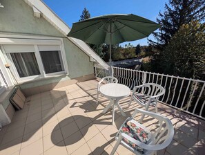 Un balcon avec un sol carrelé, une table blanche et deux chaises, un parasol vert et une balustrade blanche.