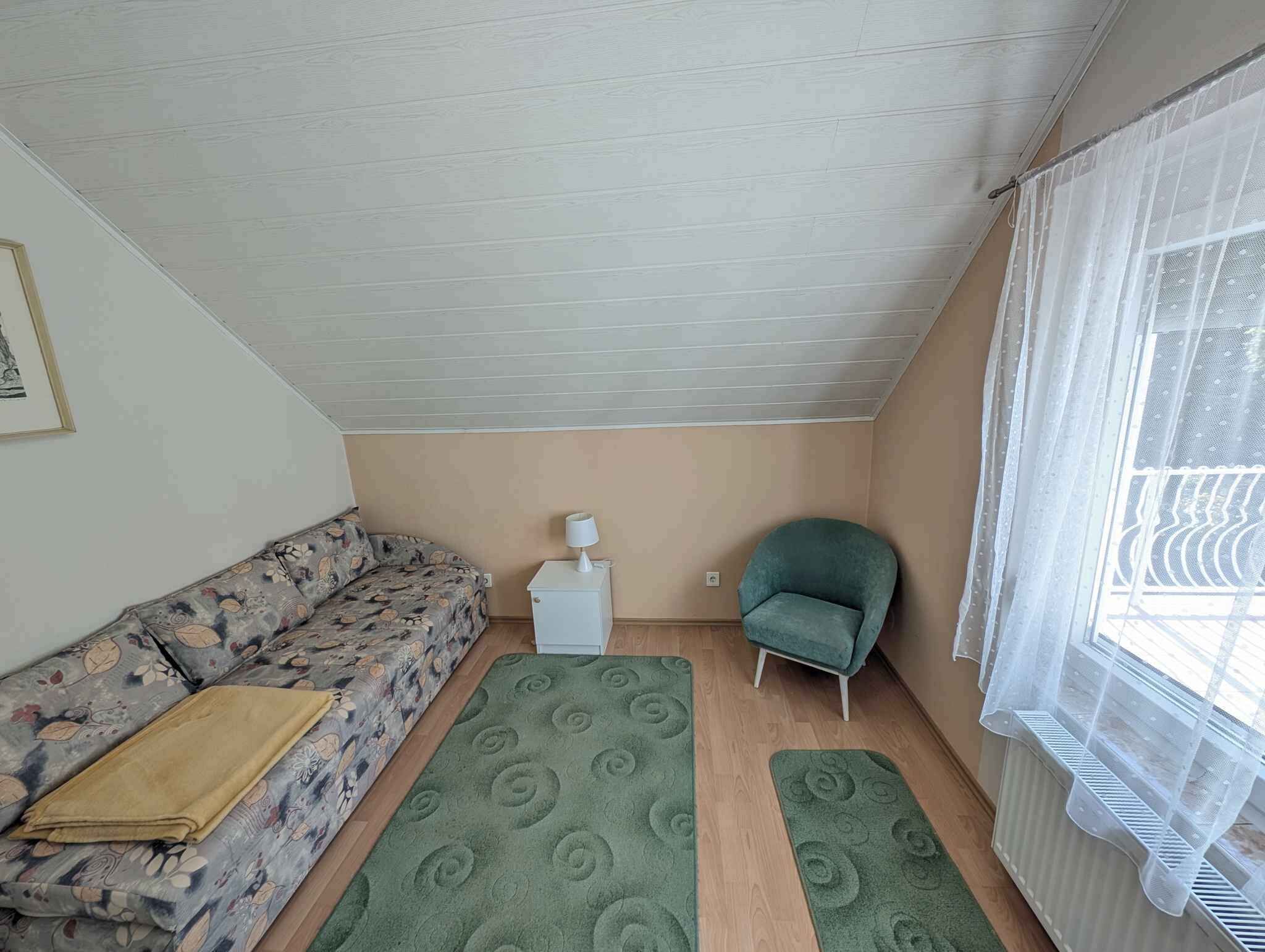 A small, cozy room with a floral sofa, a green rug, a small white table with a lamp, a teal armchair, and a window with sheer curtains.