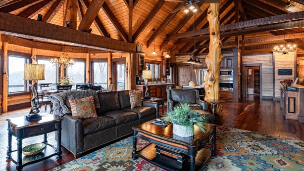A spacious living area with a high ceiling, wooden beams, a fireplace, a large sofa, a coffee table with plants, and a patterned rug. - The Sanctuary-Pet Friendly Luxury Cabin Rental, Sits on Top of the Mountain, High Speed Internet, Hot Tub, 30 Minutes to Great Smoky Mountains, in Nantahala NC (Topton)