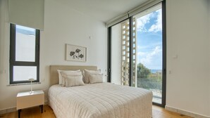 A bedroom with a large bed, a bedside table, a window with blinds, and a view of the outdoors.