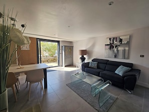 Een moderne woonkamer met een glazen salontafel, een donkere bank en een eethoek met stoelen en een tafel.