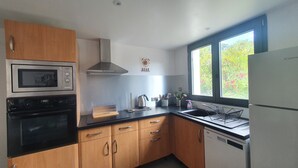A modern kitchen with wooden cabinets, a built-in oven, a microwave, a stainless steel range hood, and a dishwasher. There is a window providing natural light and a view of greenery outside. - Les Lauriers Roses: Heated pool, unspoilt nature (Saint-Vincent-de-Barrès)