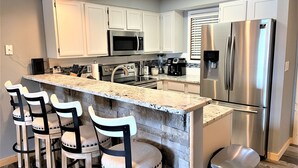 Una cocina moderna con armarios blancos, electrodomésticos de acero inoxidable, encimera de granito y taburetes de bar.
