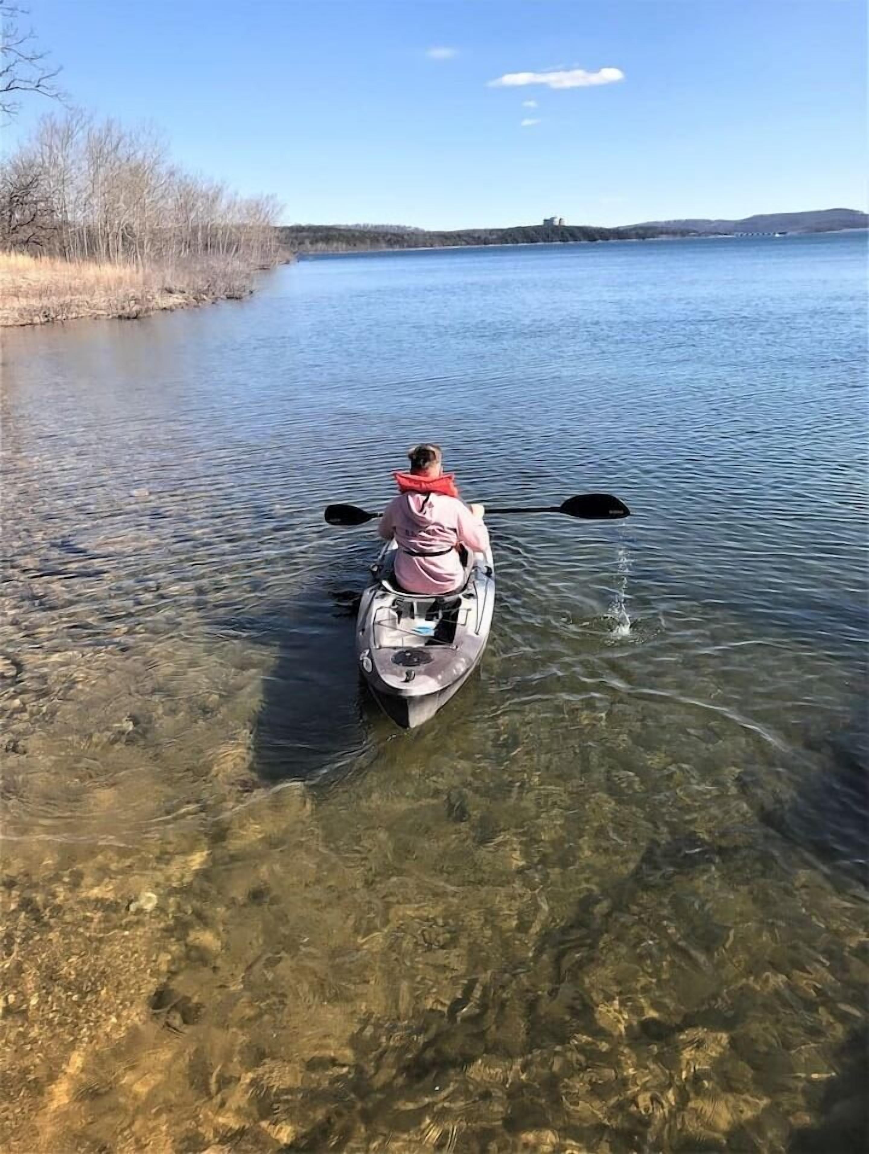 Una persona rema en kayak en un lago tranquilo, con vegetación visible en la orilla y colinas a lo lejos.