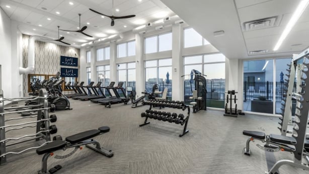 Un gimnasio amplio con cintas de correr, mancuernas y máquinas de pesas, ventanas grandes y un techo alto con ventiladores.