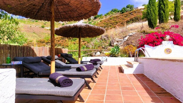 A sunlit outdoor lounge area with lounge chairs, a thatched umbrella, and a white wall with a decorative emblem. - Casa 3/Andalusian architecture in a beautiful green oasis/1 km from El Borge (El Borge)
