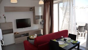 A modern living room with a red sofa, a dining table set with plates and cutlery, a flat-screen TV, and a fan. - Pilgrim's Rest Penthouse (Gzira) (Gzira)