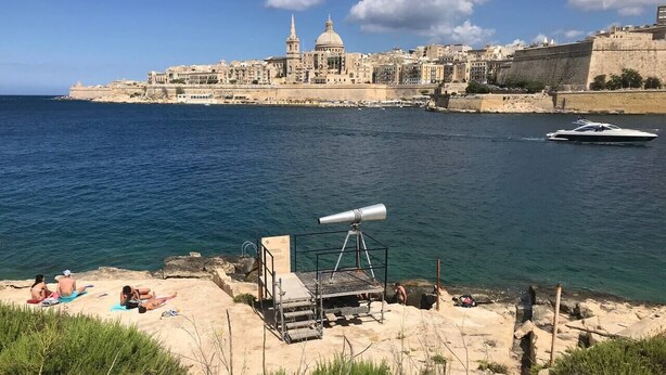 A coastal area with a telescope on a sandy beach, a clear view of a historic city with a prominent dome and tower in the background, and a boat on the water. - Pilgrim's Rest Penthouse (Gzira) (Gzira)