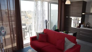 A modern living room with a red sofa, a dining table with chairs, a kitchen area with a stove, and a view of the city through large windows. - Pilgrim's Rest Penthouse (Gzira) (Gzira)