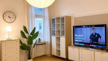 Ein modernes Wohnzimmer mit Fernseher, der eine Sendung zeigt, einem Essbereich mit Tisch und Stühlen, einer Zimmerpflanze und einem Bücherregal.