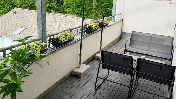 Un balcón al aire libre con una mesa, sillas, un parasol y plantas en macetas.