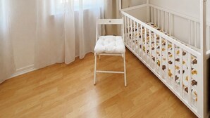 Una cuna blanca sobre piso de madera, una silla blanca y una ventana con cortinas de encaje.