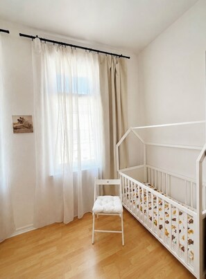 A white crib with a wooden floor, a white chair, and a window with sheer curtains.