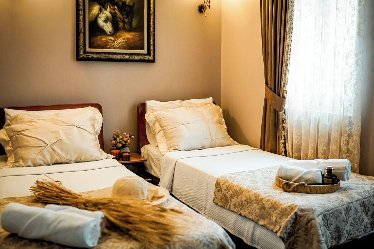 A hotel room with two beds, a painting on the wall, a nightstand with a vase of flowers, and a window with curtains.