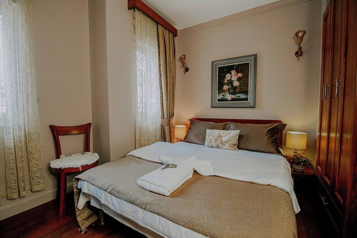 A neatly made bed with a white and beige duvet, a wooden chair with a white cushion, a painting of flowers on the wall, and a wooden wardrobe.
