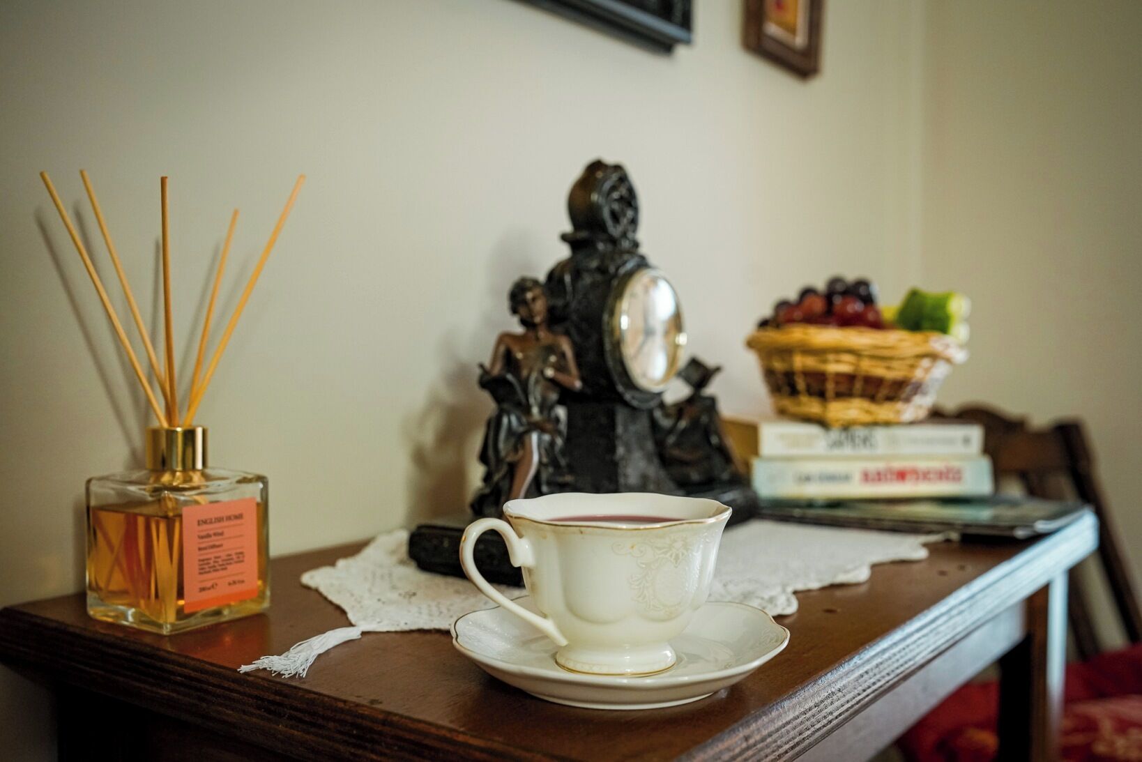 Une table avec une tasse et une soucoupe blanches, un flacon de parfum d’ambiance de la marque anglaise English Home, une figurine et un panier de fruits.