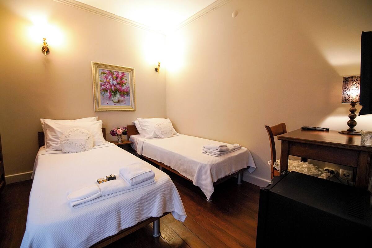 A hotel room with two single beds, a desk, and a chair. There are towels on the beds and a painting on the wall.