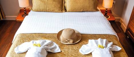 Un letto rifatto con un copriletto bianco e due cuscini dorati, due lampade da comodino e due asciugamani con un fiore giallo su un tenda dorata.