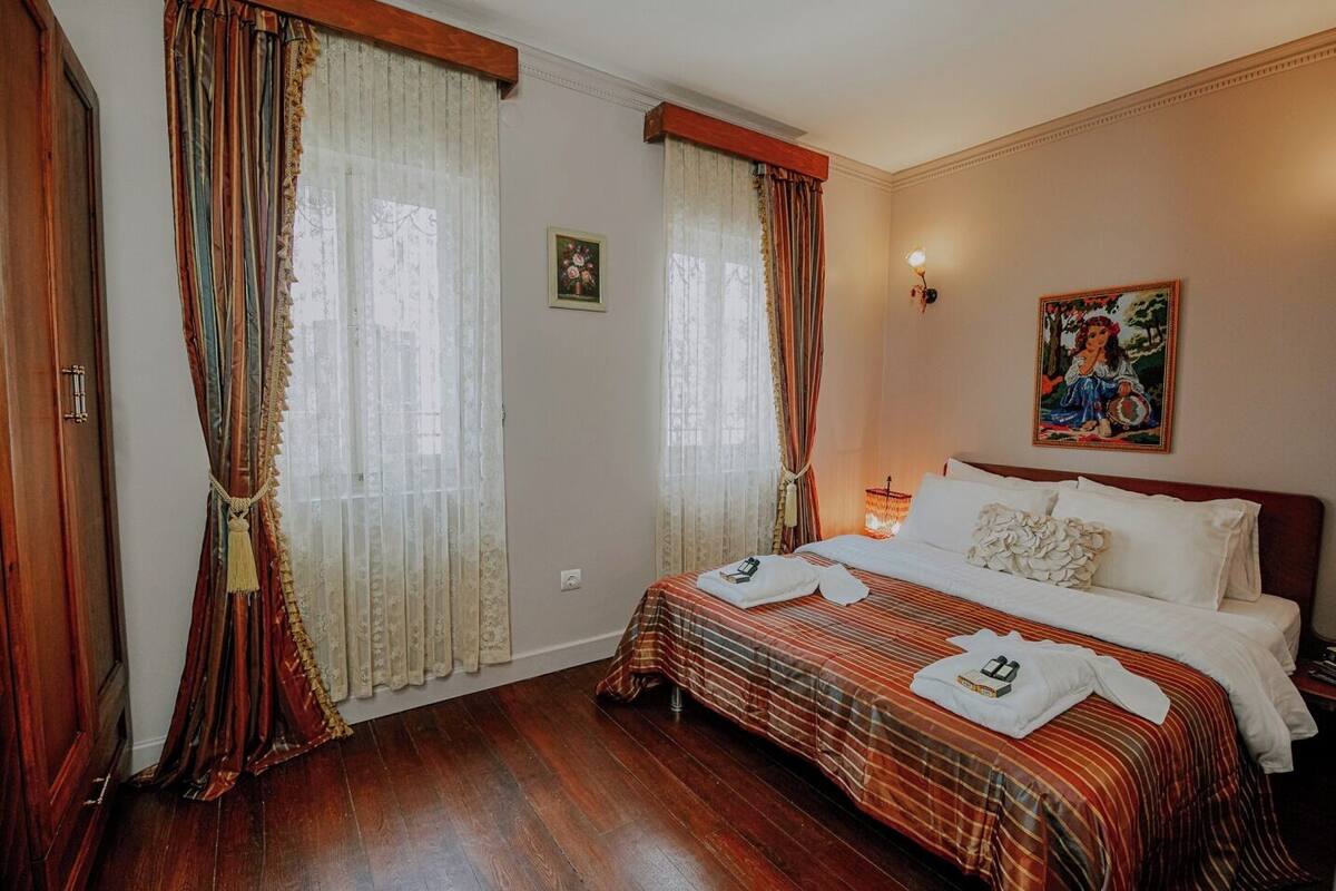 A hotel room with a large bed, two windows with curtains, a painting on the wall, and a wooden floor.