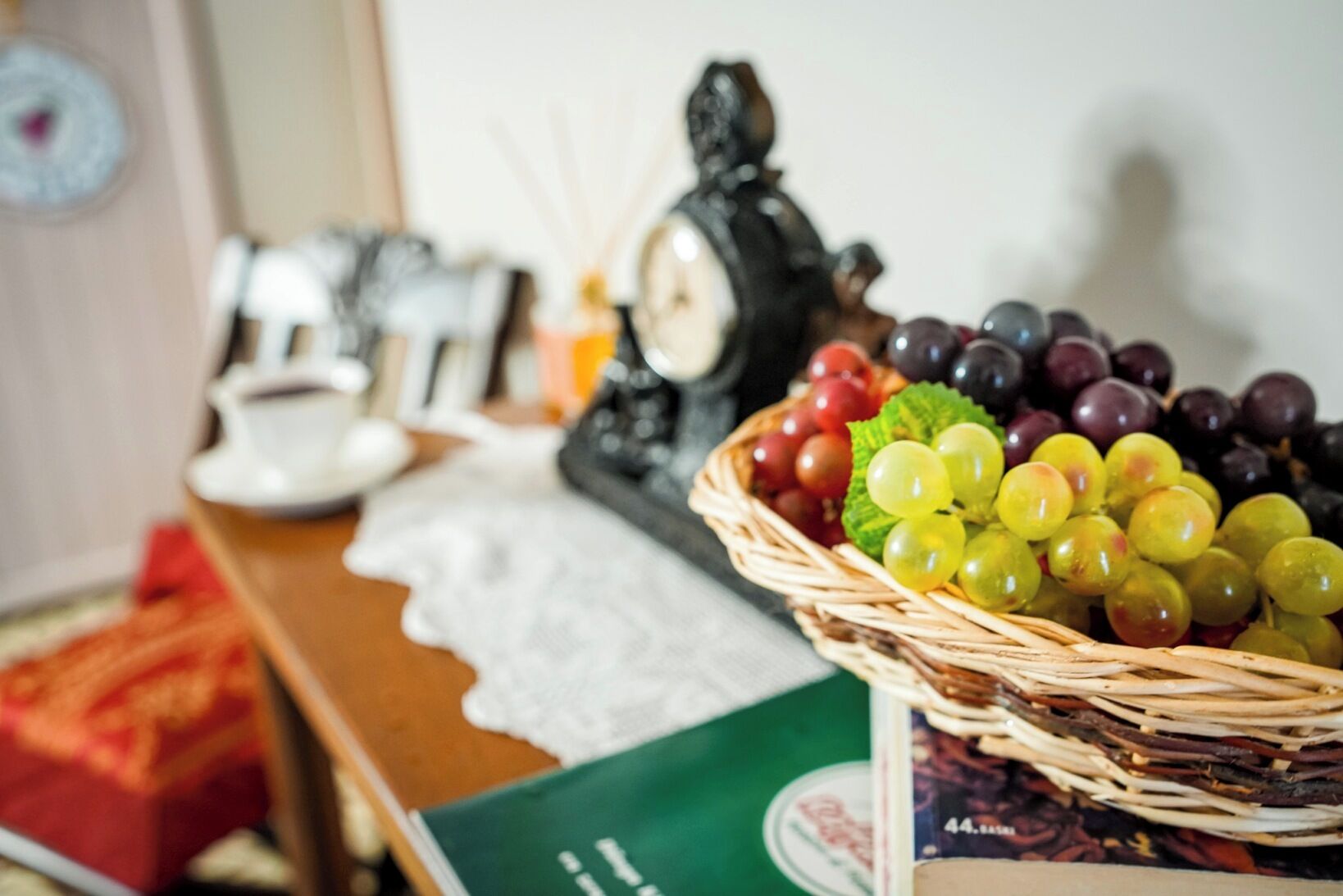 Una cesta de mimbre llena de uvas de varios colores, una figura decorativa de un pingüino y una tetera de cerámica blanca sobre una mesa.
