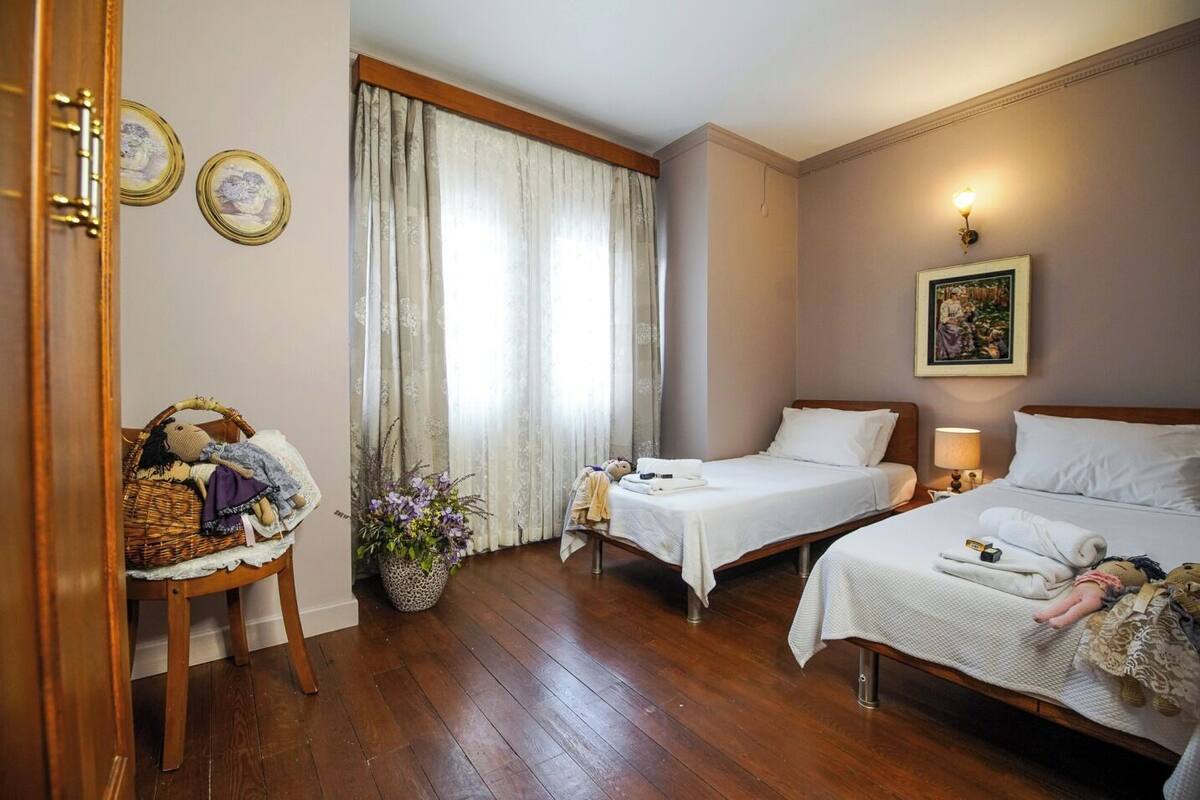 A hotel room with two single beds, a wooden floor, a large window with curtains, a wall-mounted picture, and a wicker chair with a blanket.