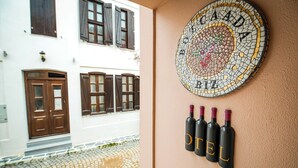 A hotel entrance with a cobblestone path, wooden doors, and windows with shutters. A mosaic artwork with the word "HOTEL" is displayed on the wall, and four wine bottles are arranged below it.