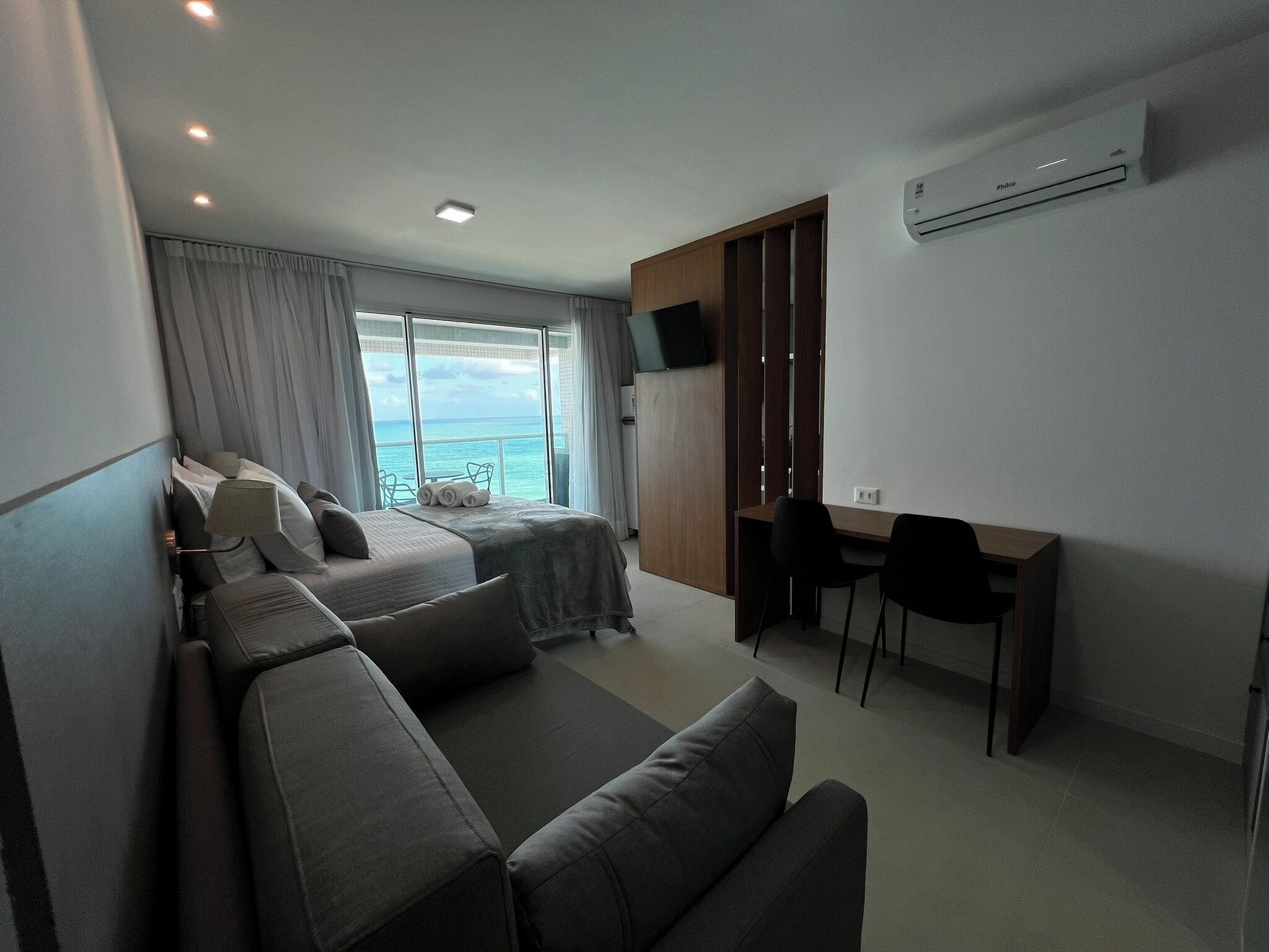 A modern hotel room with a bed, a sofa, a desk, and a view of the ocean.