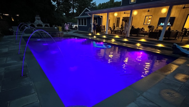 Una piscina con iluminación azul, baldosas de piedra y una zona cubierta con asientos y una televisión.
