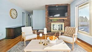 Ein Wohnzimmer mit Kamin aus Backsteinen, einem Flachbildfernseher, einer Uhr und zwei weißen Sesseln.