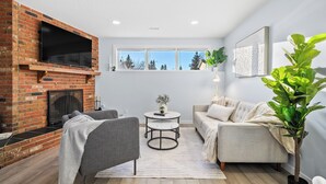 Ein Wohnzimmer mit Kamin aus Ziegelsteinen, einem Flachbildfernseher, einem grauen Sofa, einem runden Couchtisch und einer großen Topfpflanze.