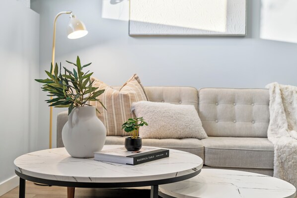 A modern living room with a round coffee table, a white vase with green plants, a book titled 'Magnolia Table', a sofa with a white throw blanket, and a floor lamp.