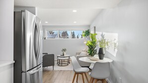 Eine moderne Küche mit einem Edelstahl-Kühlschrank, einem Essbereich mit rundem Tisch und Stühlen sowie Blick auf Bäume durch große Fenster.