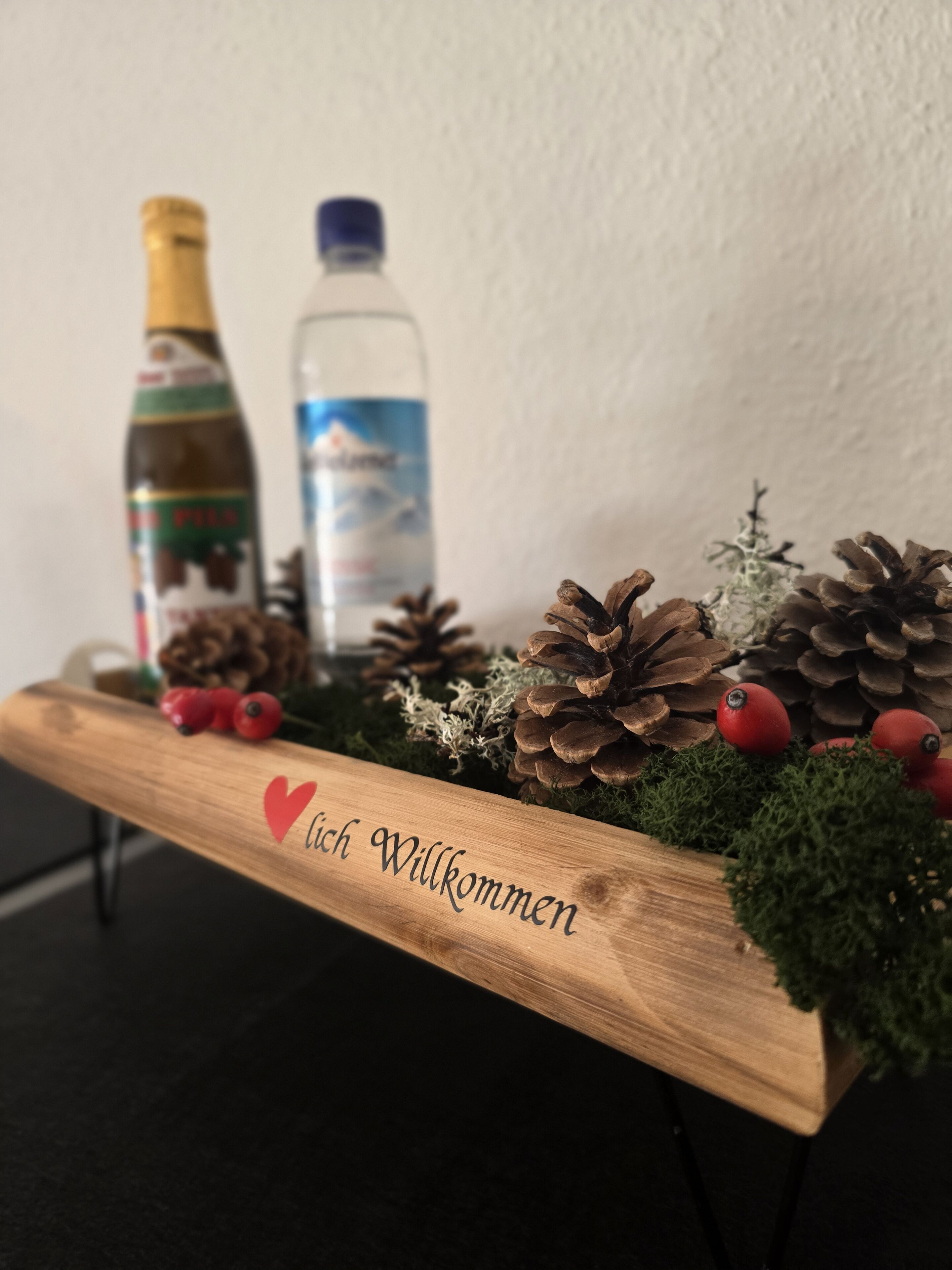 Een houten plankje met een 'Willkommen'-bord, denne-appels, rode bessen en twee flessen.