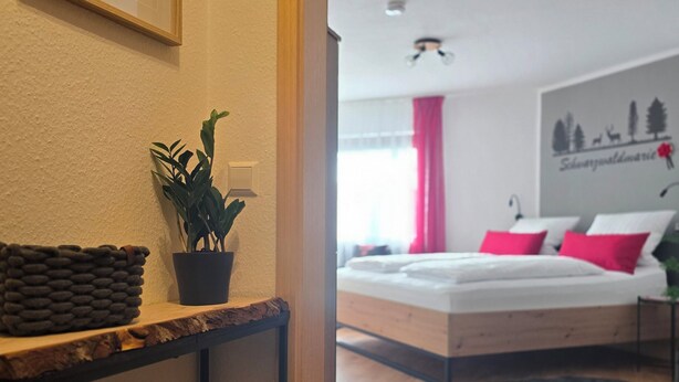 A modern hotel room with a wooden floor, a bed with pink and white bedding, a bedside table with a plant, and a framed picture on the wall. - Schwarzwaldmarie Titisee bathtub kitchen balcony underground parking near the lake! (Titisee-Neustadt)